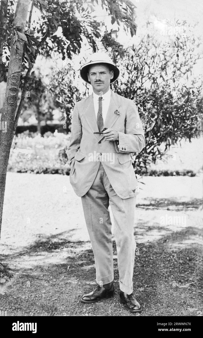 Posed photograph of an unidentified European man in a garden under some trees. He is smartly dressed and wearing a pith helmet, holding a pipe in his left hand with right hand in trouser pocket. Number 68, possibly a house number, is cut into the bark of the tree to his left. Stock Photo