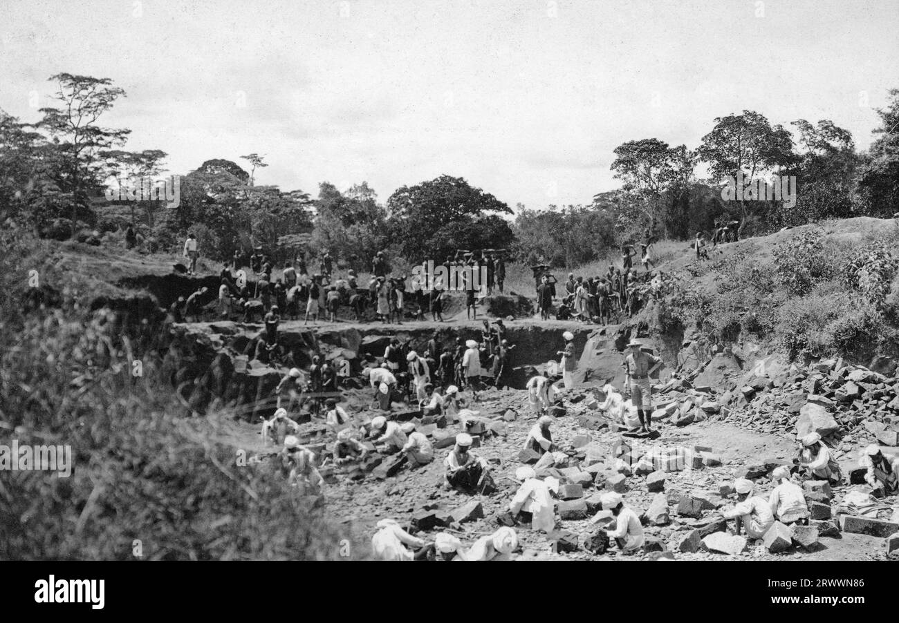 Photograph of a shallow stone quarry with hundreds of workers using hand labour to break the stone and porters carrying away the best on their heads. In the foreground are mainly Sikh men cutting the blocks, and African men behind are working as labourers. A European man in the centre of the image is acting as overseer. Original manuscript caption: Stone Quarry nr Pangani Village Nairobi 1913. Stock Photo