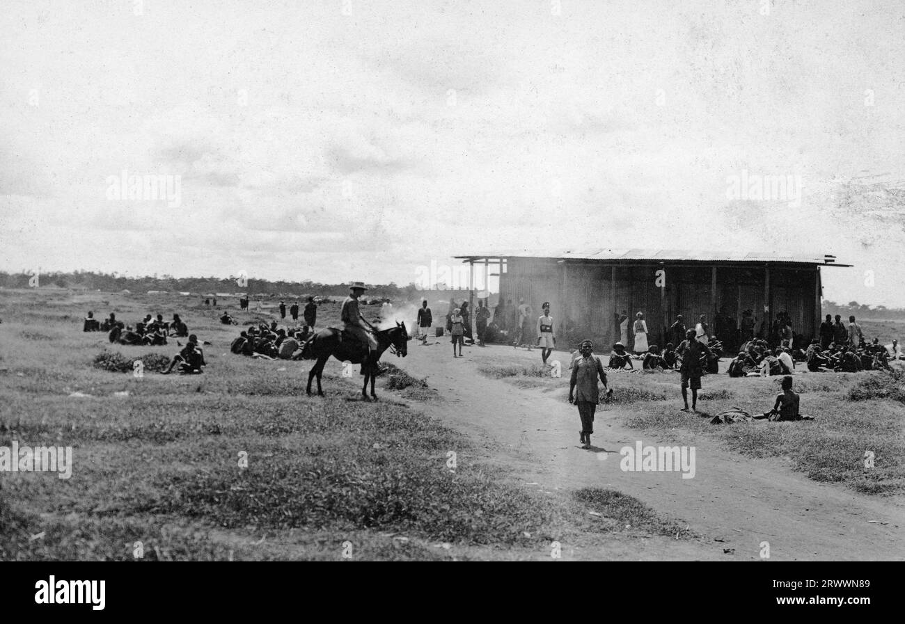 A large building made from corrugated iron around a wooden framework is surrounded by a large number of people, mainly African men, sitting or standing around. The majority are talking in groups. Also visible in the shot is a Kenyan policeman and a European man on horseback. Open grassland surrounds the market. Original manuscript caption: Old Native Market. Nairobi 1912. Stock Photo