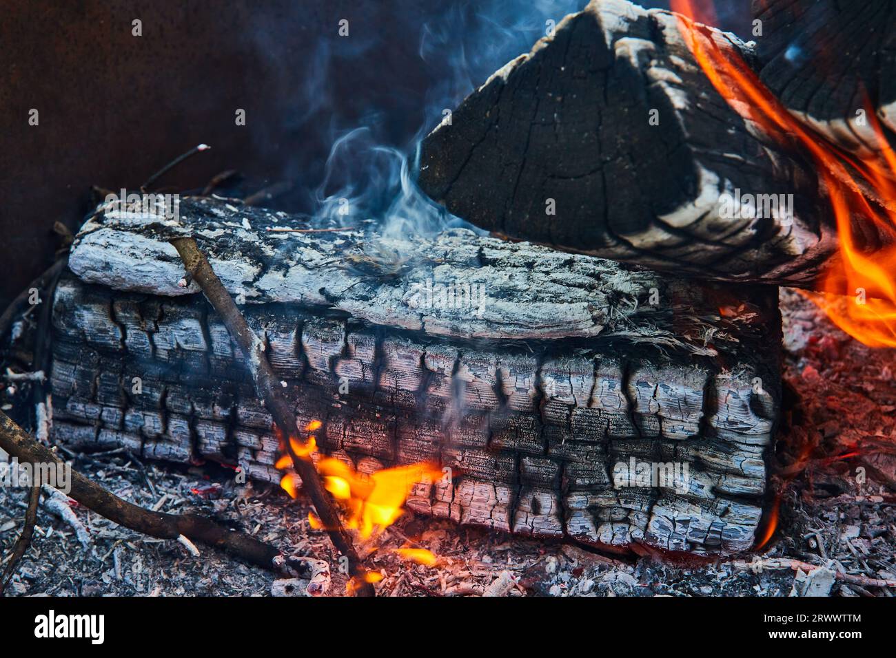 Burnt logs with orange and yellow flames and smoke rising up close up