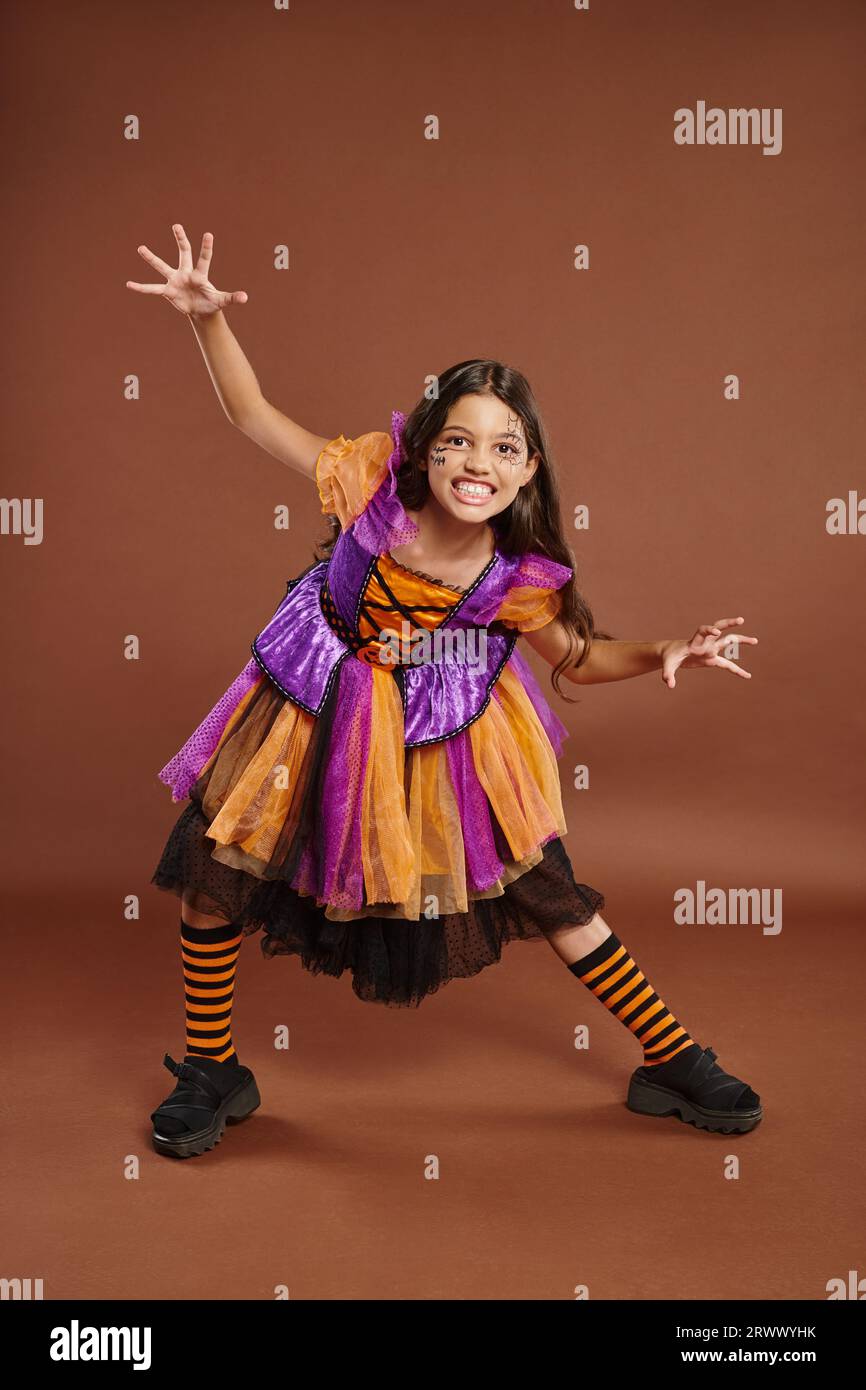 spooky girl in Halloween witch costume growling and gesturing on brown backdrop, full length Stock Photo