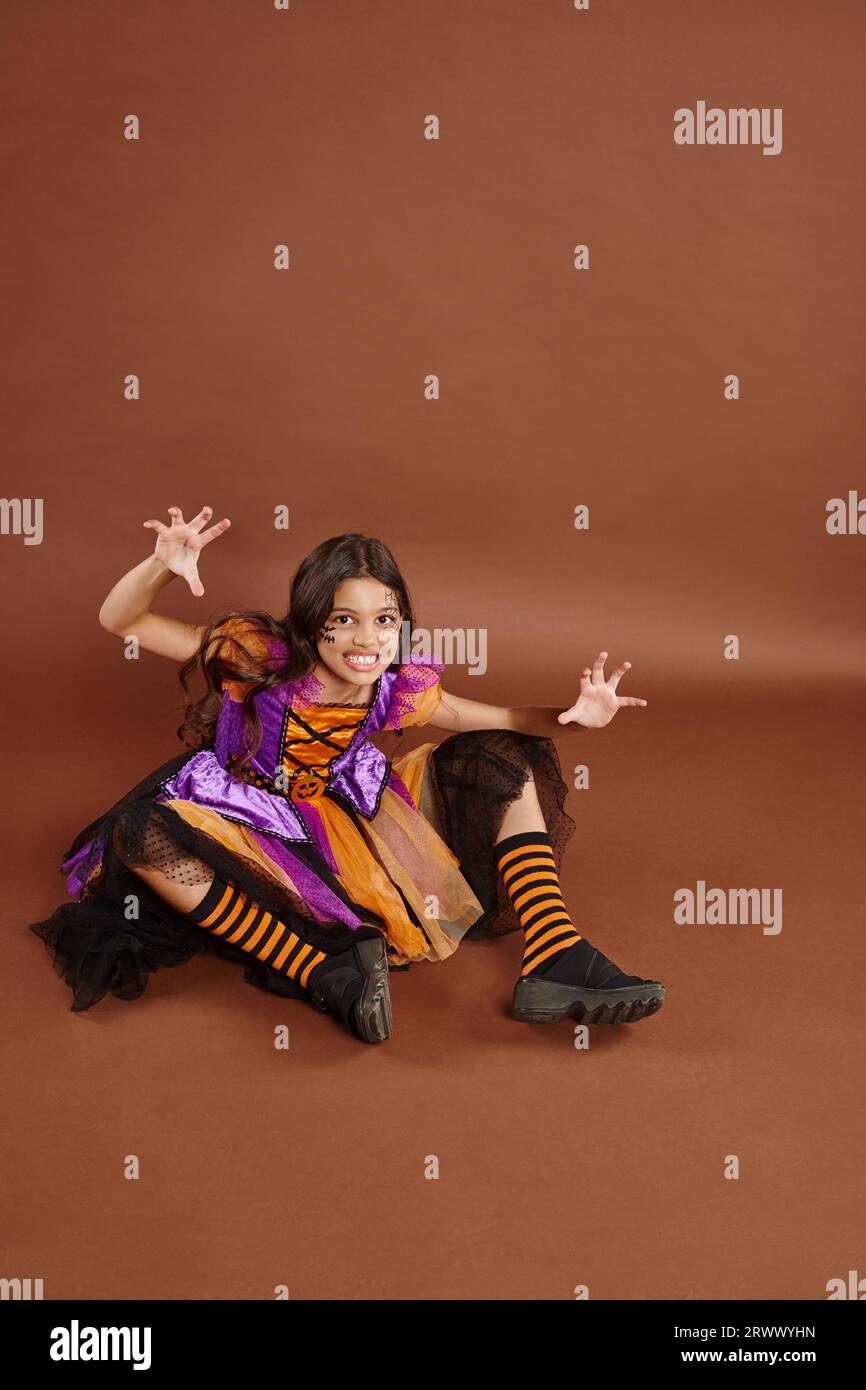 spooky girl in Halloween witch costume sitting and growling on brown backdrop, October 31 Stock Photo