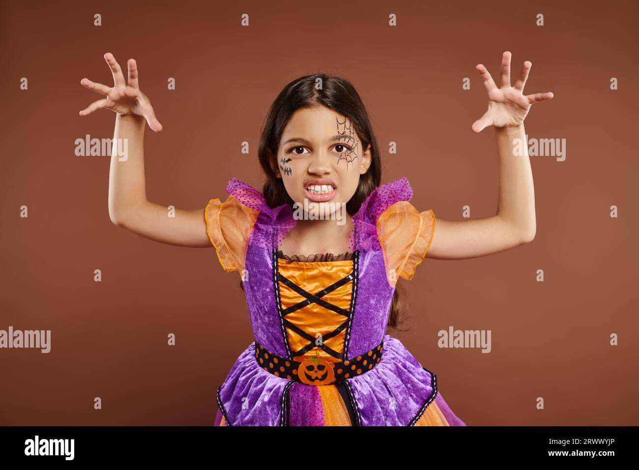 spooky child in Halloween costume with spiderweb makeup growling and gesturing on brown backdrop Stock Photo