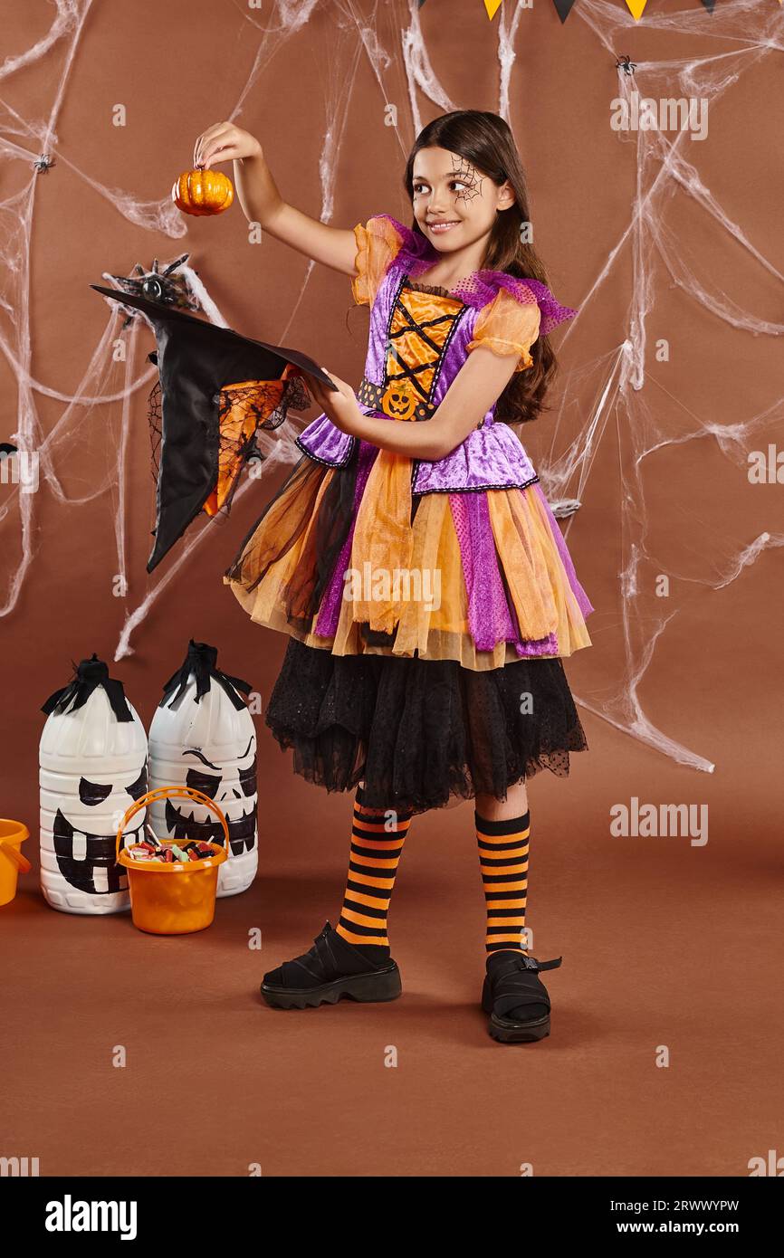 happy child in Halloween witch costume holding pumpkin near pointed hat on brown background Stock Photo