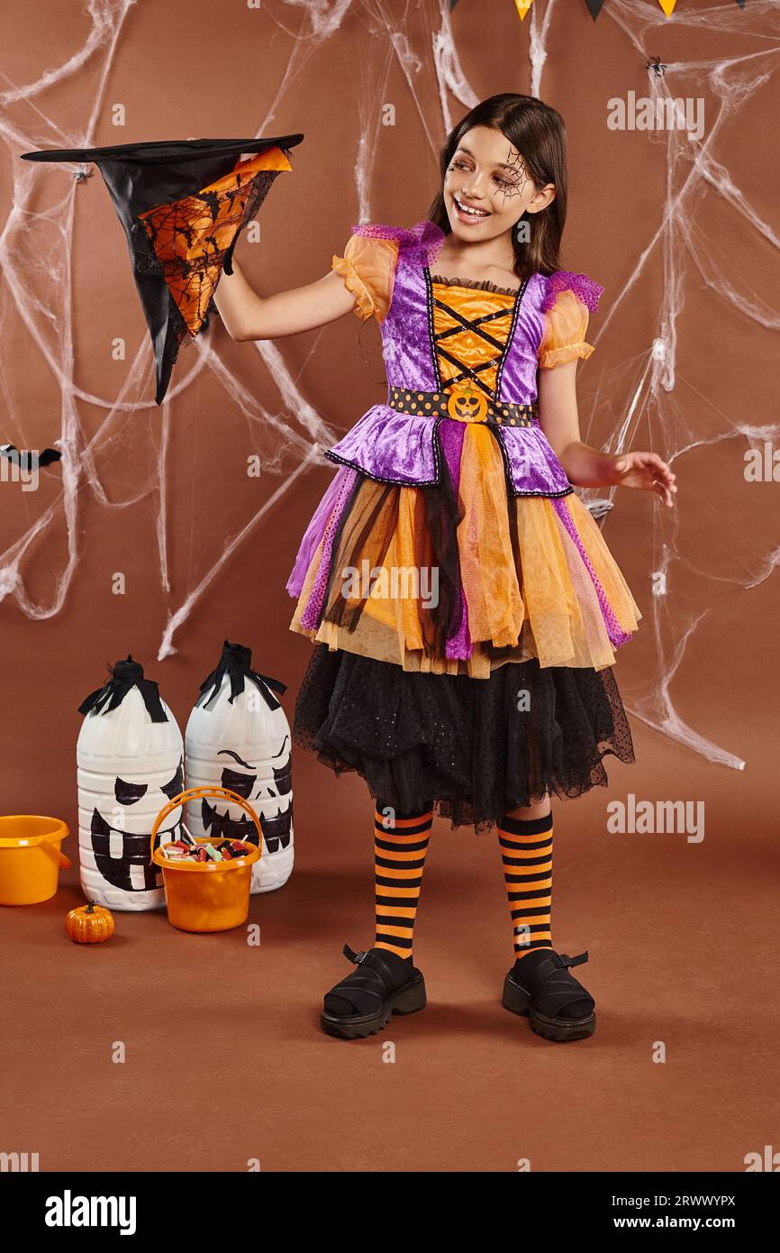 cheerful child in Halloween witch costume holding pointed hat upside down on brown background Stock Photo