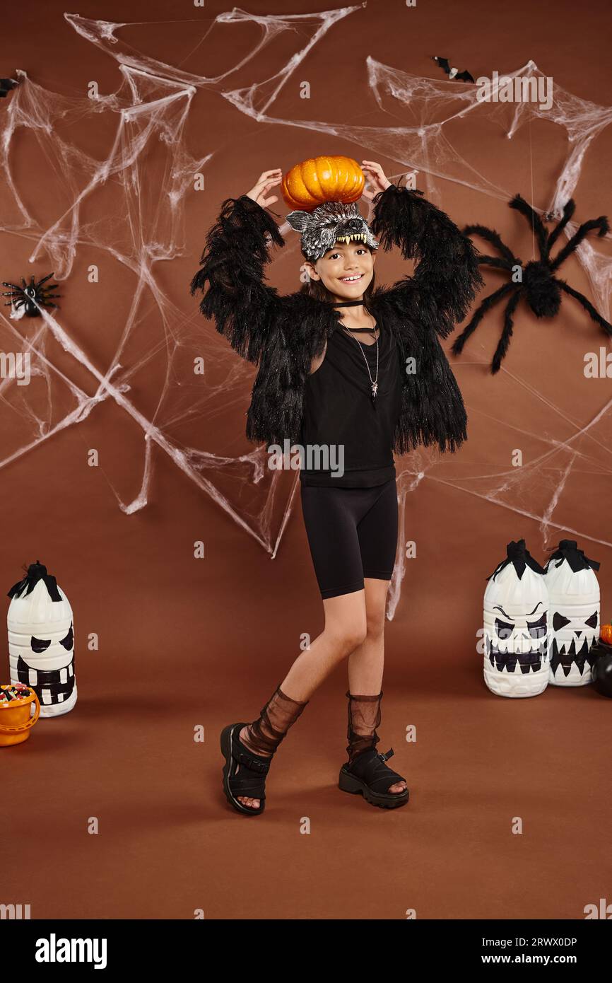 happy preteen girl holds pumpkin on her head with hands, brown backdrop