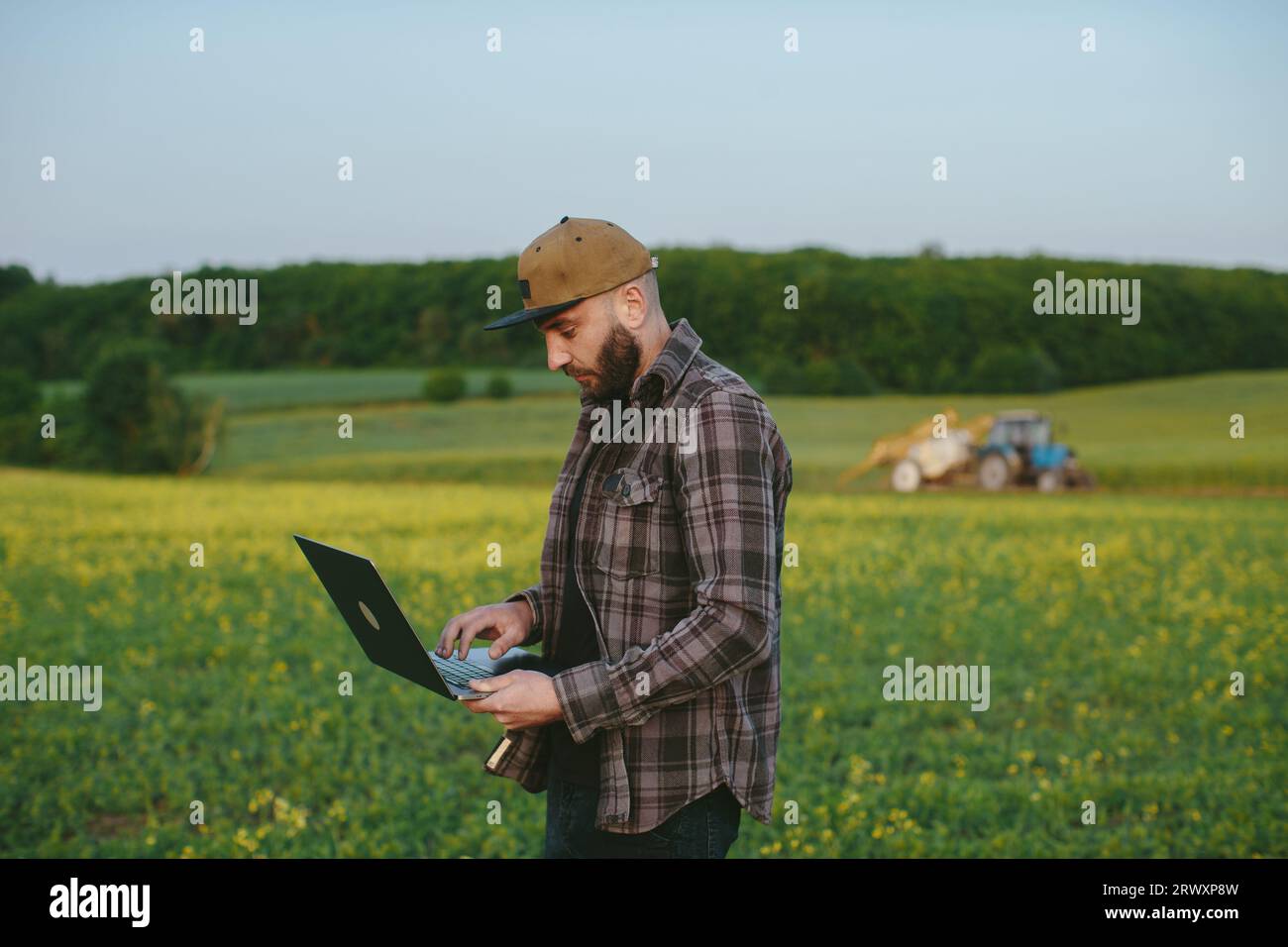 Caucasian young male farmer in hat standing in field and typing on keyboard of laptop computer. Attractive man using device in margin in summer and br Stock Photo