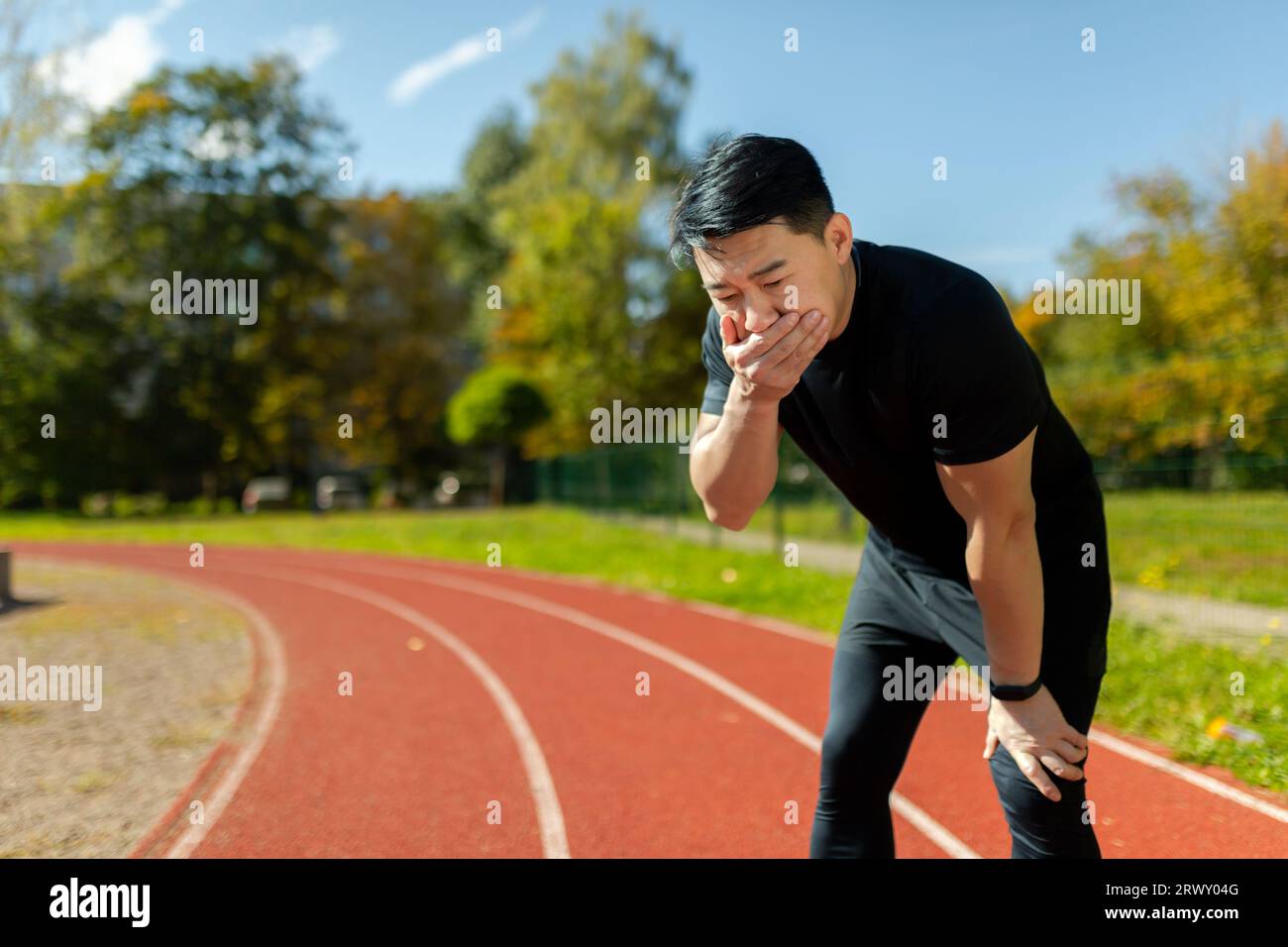 Asian young man doing sports at the stadium. He stands bent over on the treadmill, covers his mouth with his hand, feels very nauseous. Stock Photo