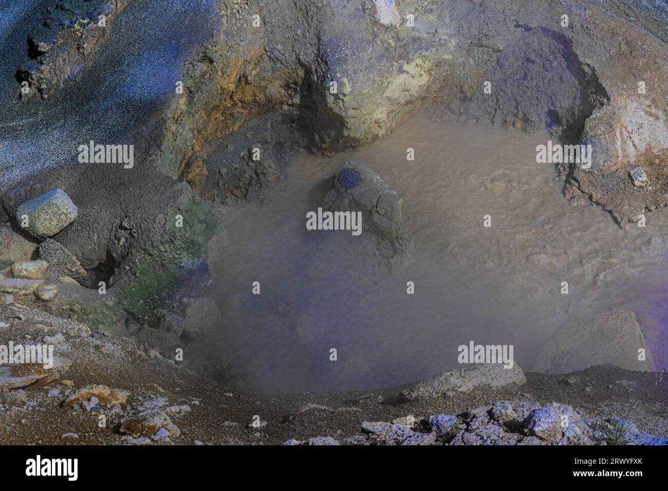 Top view shows mud surface with volcanic mineral rock & sediment in colors. Ground slopes to watery muddy pool with bubbling muck in Yellowstone Park. Stock Photo