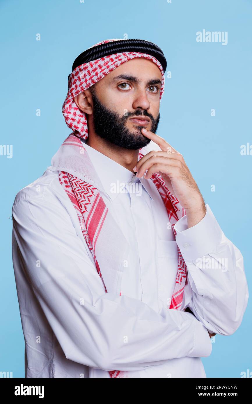 Thoughtful muslim man wearing thobe and checkered headscarf standing in deep thoughts with pensive emotion. Questioned arab person posing in contemplation on blue background Stock Photo