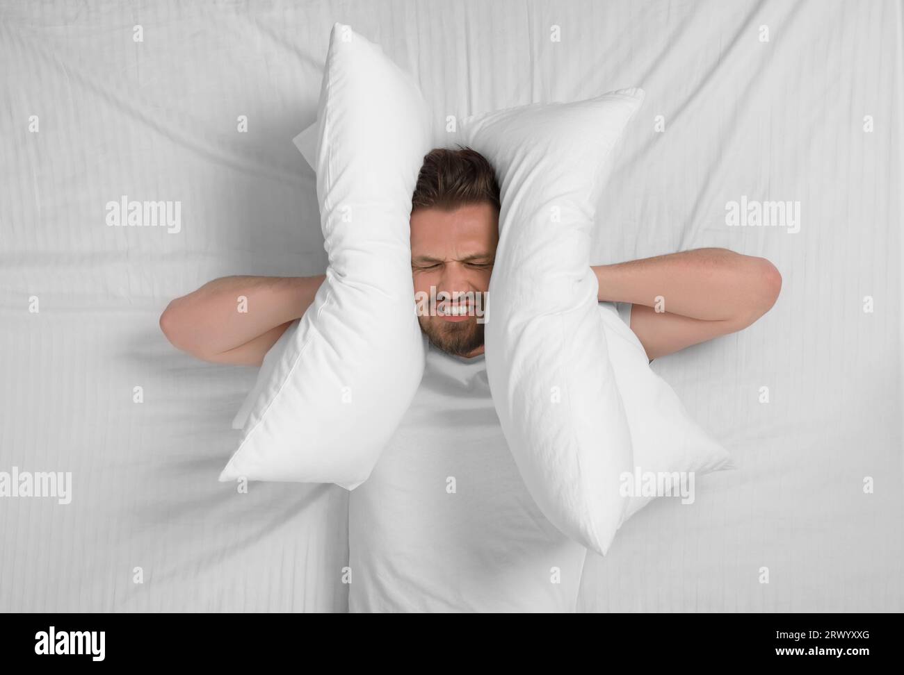 Irritated man covering his ears with pillows in bed, top view. Insomnia