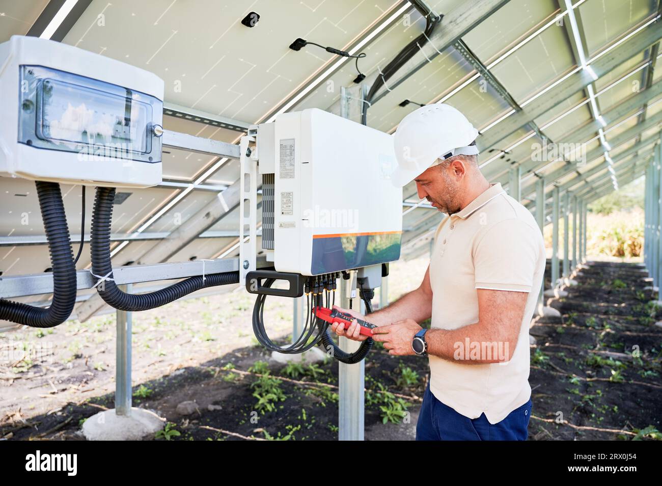 Good worker checking solar panel voltage. Turning on solar system. Man in helmet with current clamps in hands on back side of battery. Stock Photo