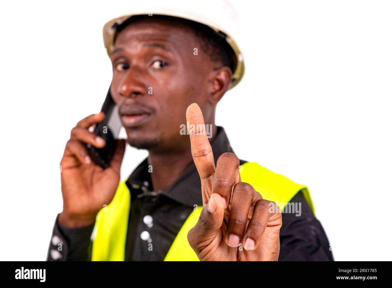 young busy engineer man wearing green vest and hard hat and talking on mobile phone while showing waiting gesture with raised index finger. Stock Photo
