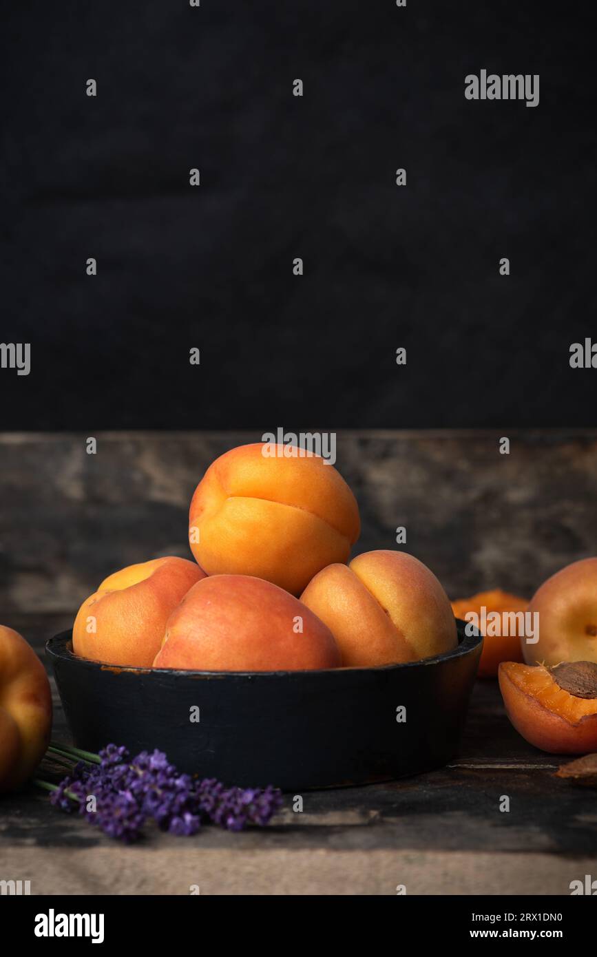 Fresh, ripe, juicy apricots in a black bowl on a wooden table. Whole fruit, fruit around a bowl, half apricot and a purple flower on the table Stock Photo