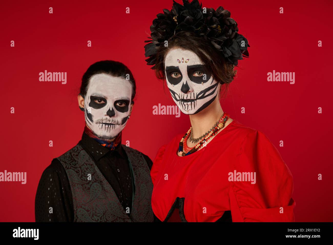 eerie couple in traditional catrina makeup looking at camera on red, dia de los muertos celebration Stock Photo