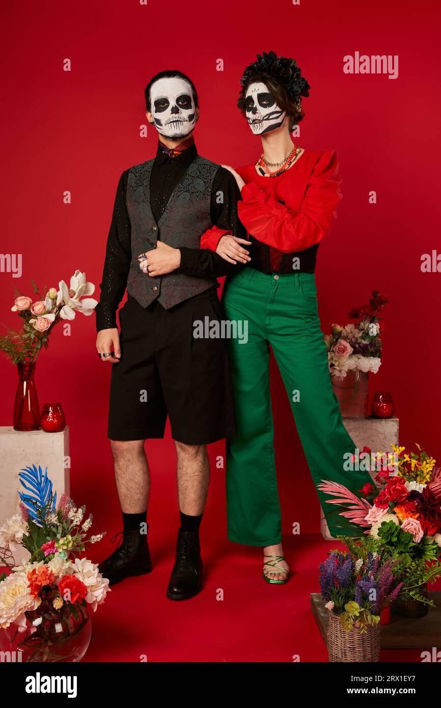 full length of couple in skull makeup near traditional Day of Dead ofrenda with flowers on red Stock Photo