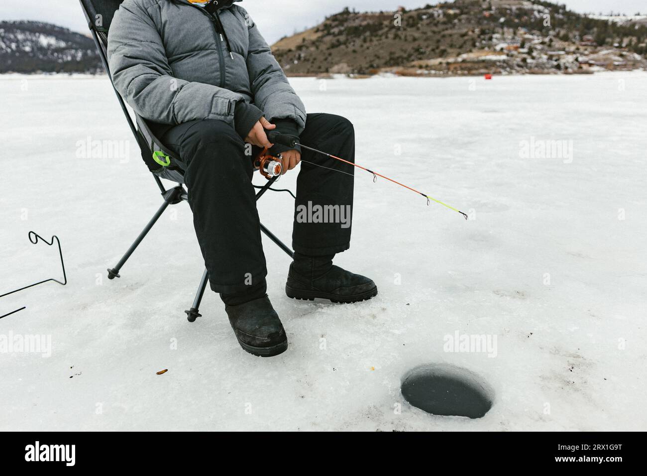 Boy sits on frozen lake fishing in ice hole in winter in coat Stock Photo