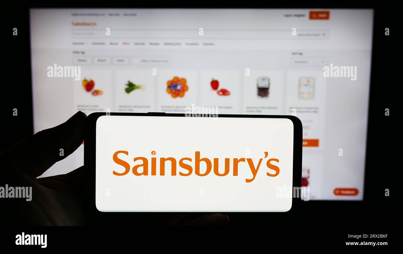 Person holding mobile phone with logo of British supermarket company J Sainsbury plc on screen in front of web page. Focus on phone display. Stock Photo