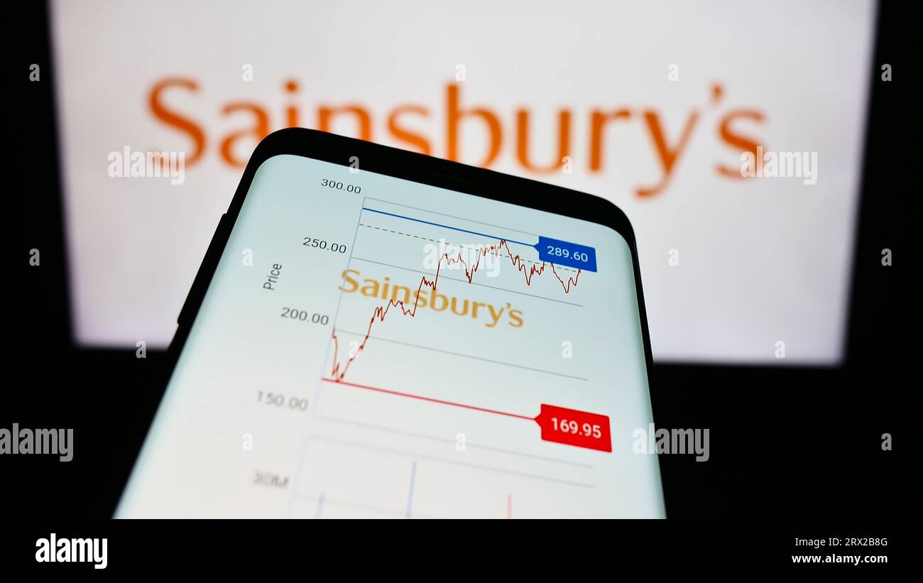 Smartphone with webpage of British supermarket company J Sainsbury plc on screen in front of business logo. Focus on top-left of phone display. Stock Photo