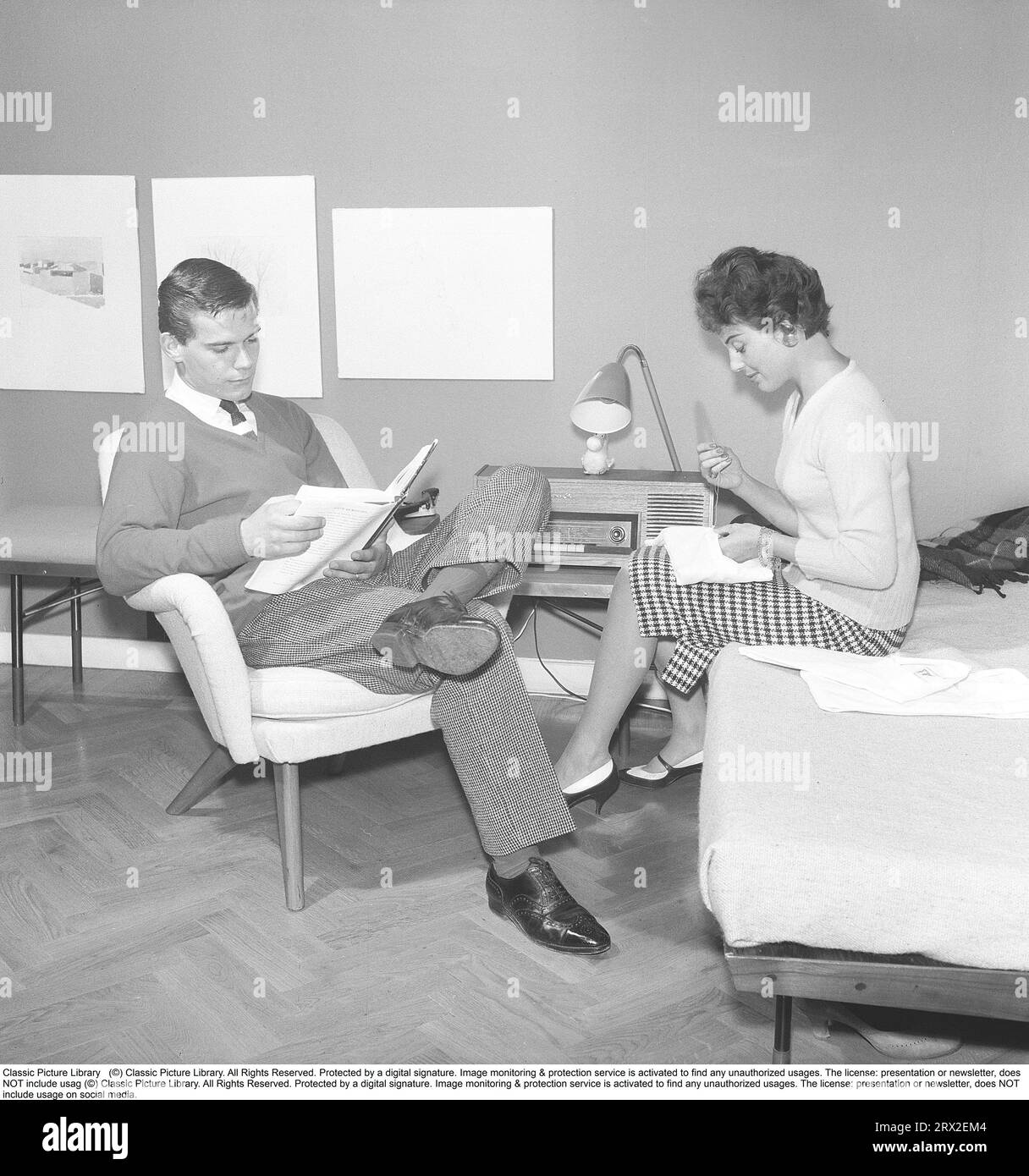 A young couple sitting together at home in the 1950s. The man sits well dressed in shirt and tie reading while the young woman sits on the bed embroidering monograms on their bedclothes and pillowcases. Maybe they recently got married and got an apartment. Sweden 1958. Kristoffersson ref CA38-10 Stock Photo