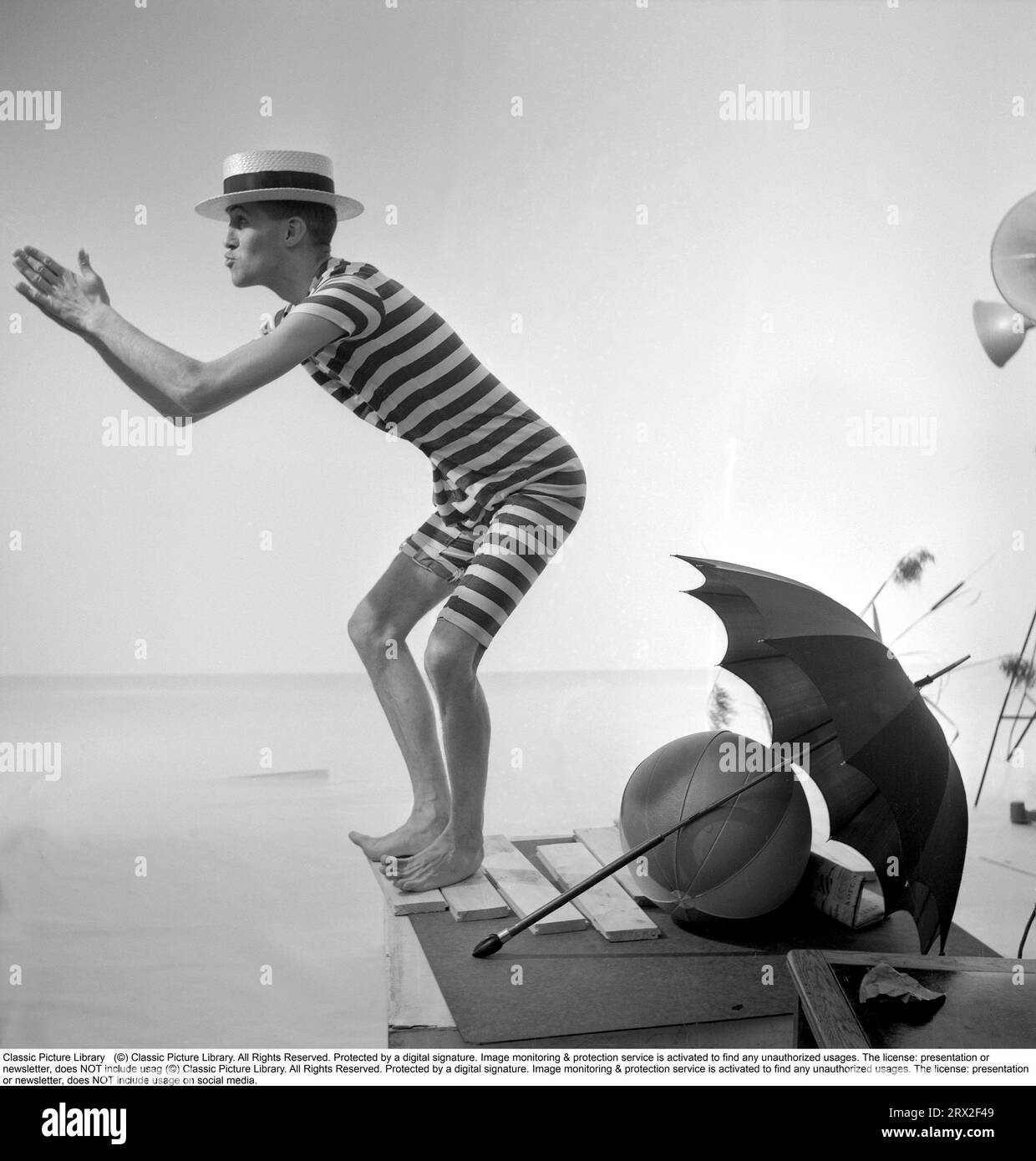 Prepared for the beach regardless of weather. The man is oddly enough dressed in a historical male bathing suit that was used in te beginning of the 20th century in a striped dark and light pattern.He wears a dixie hat and pretends to dive into the water with his hands in front of him. He is prepared for bad weather as an umbrella is in the picture. Sweden 1956 Stock Photo