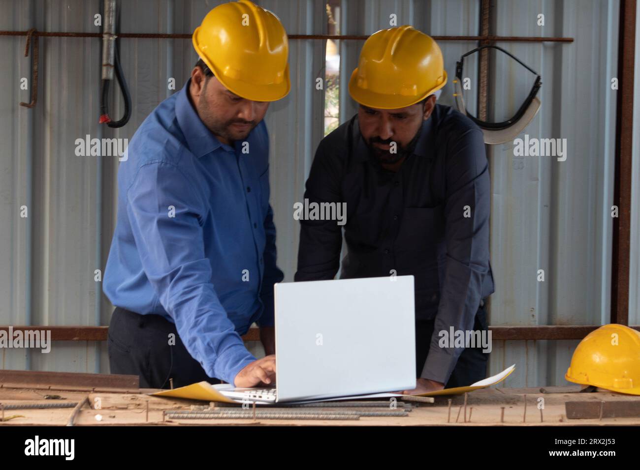 Two male construction architects using laptop and blueprint at project site. They are wearing safety helmet looking at the screen. Stock Photo