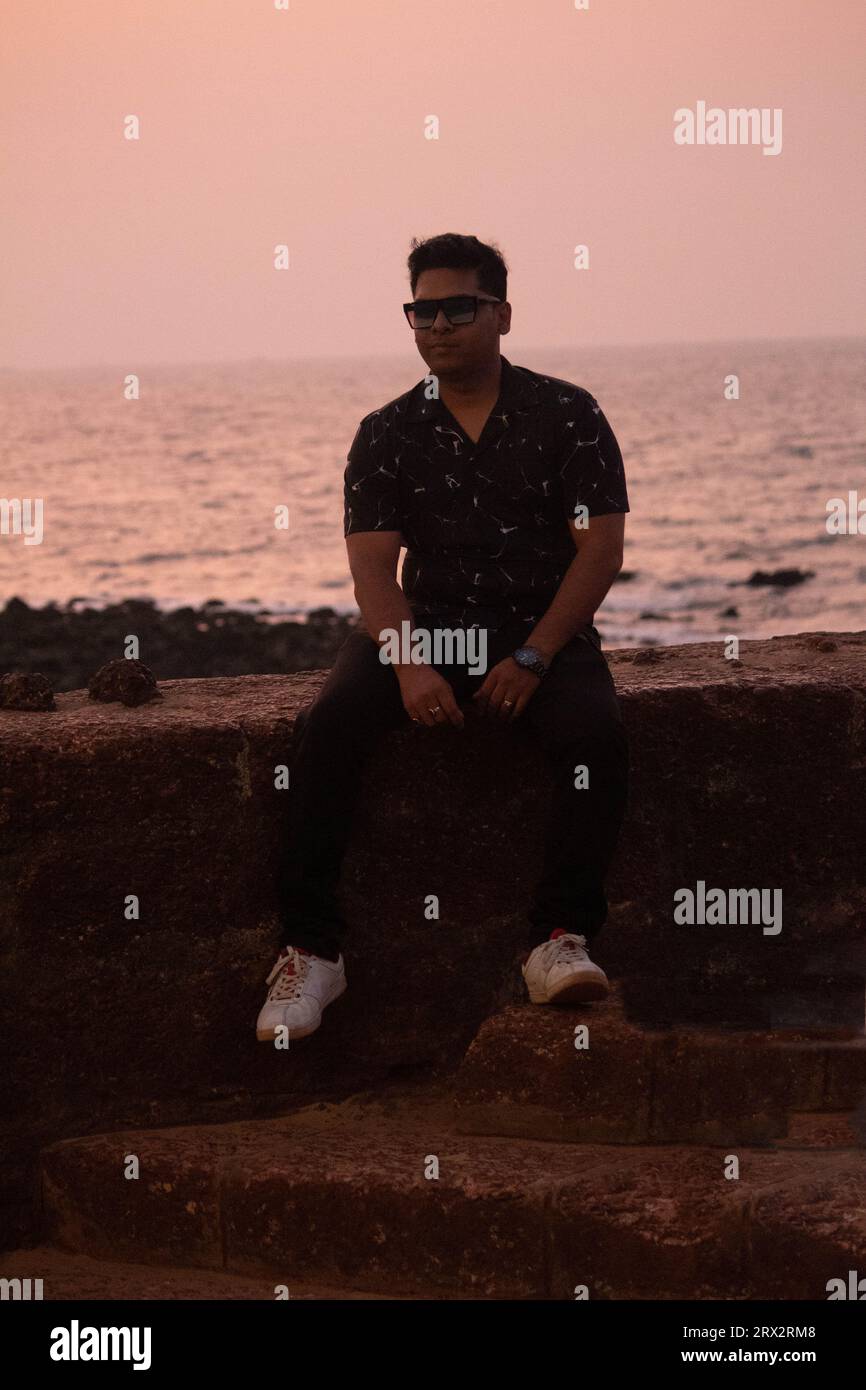 Handsome man sitting on stonewall in front of beautiful seascape during dusk. He is wearing short sleeved shirt and sunglasses Stock Photo