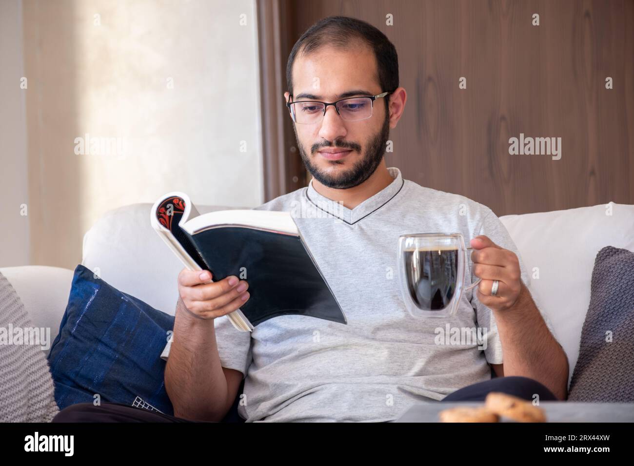 Bearded arab man drinking coffee while reading book and sitting on living room couch Stock Photo