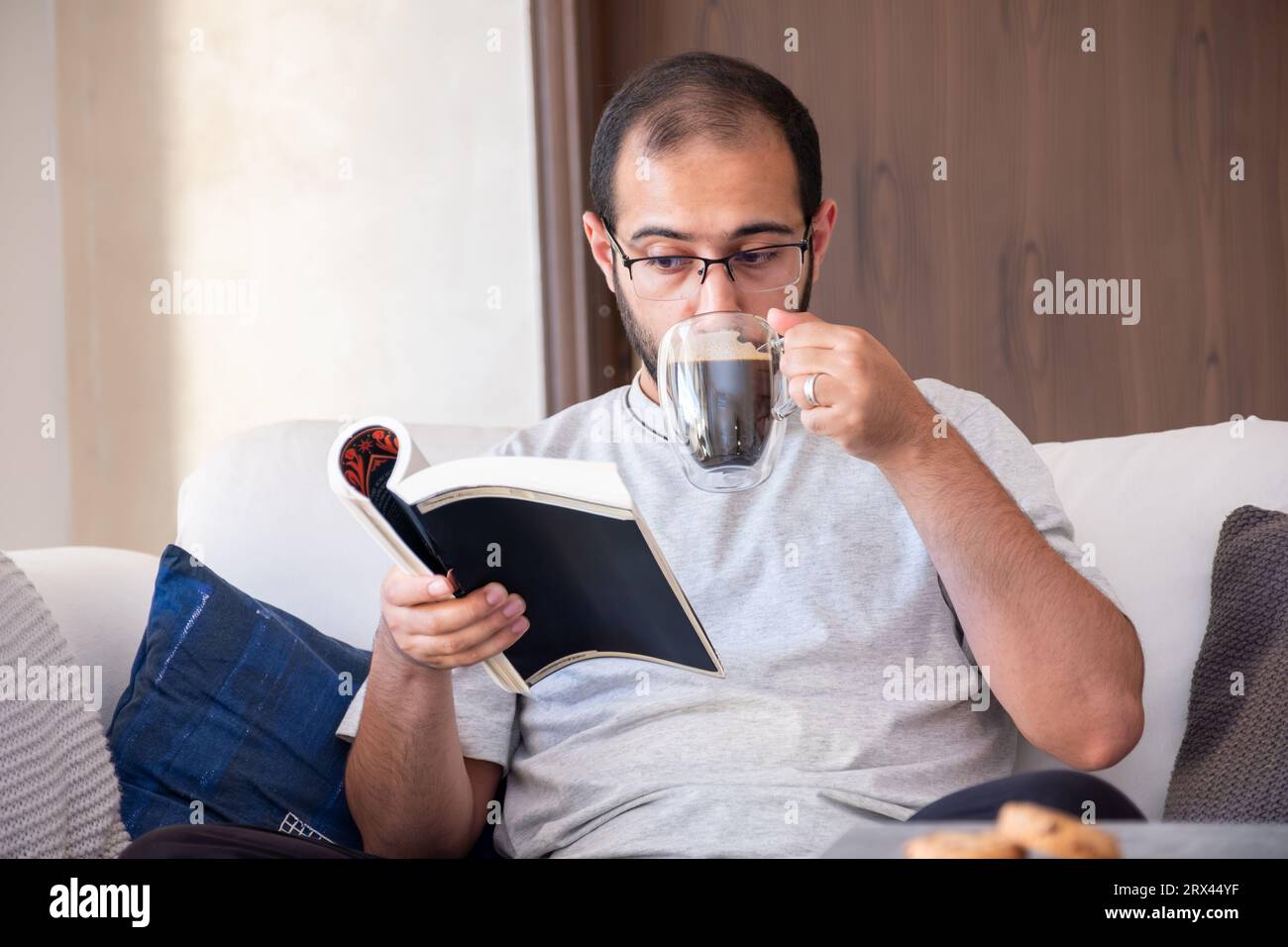 Bearded arab man drinking coffee while reading book and sitting on living room couch Stock Photo