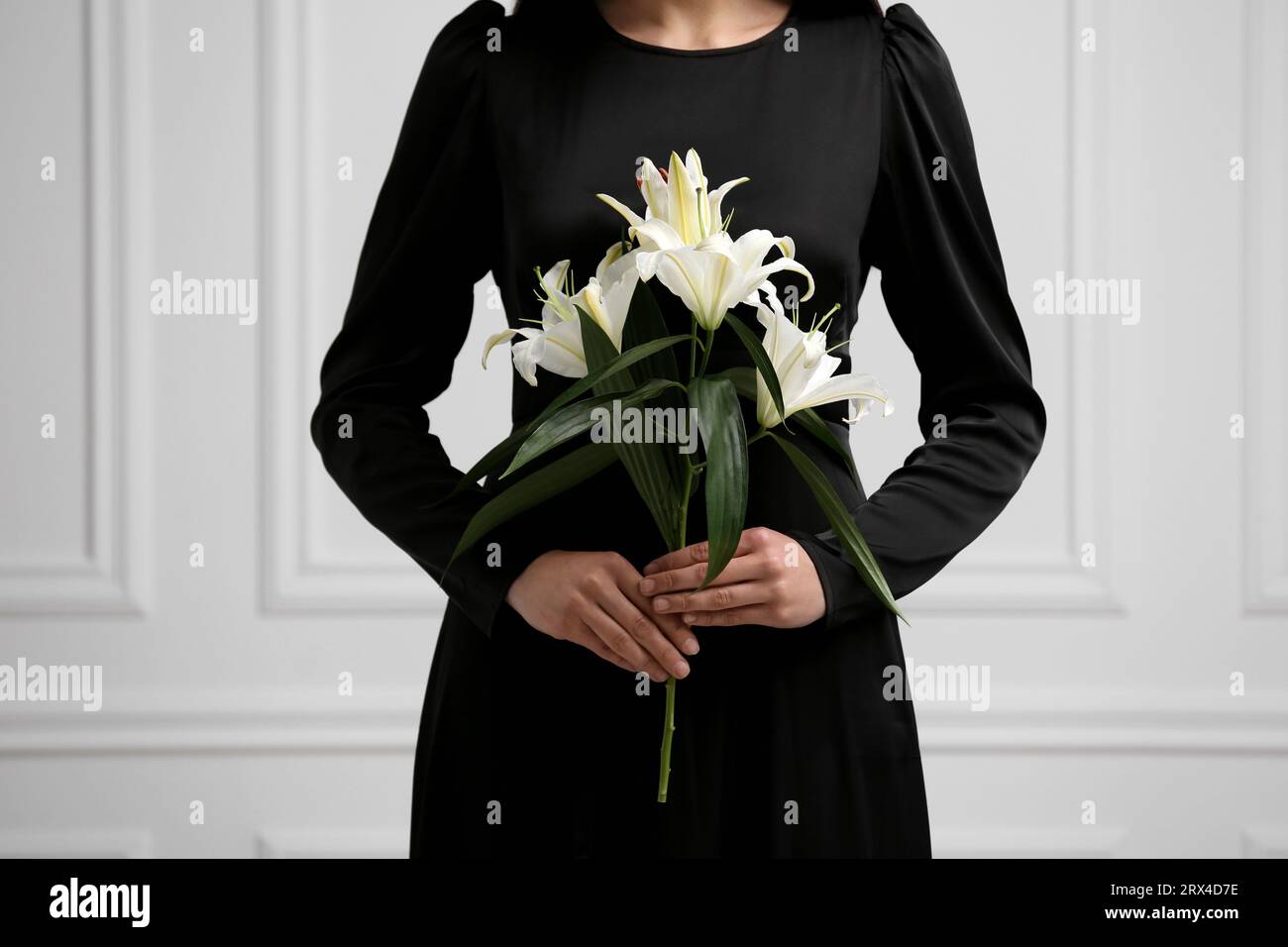 Woman with lilies near white wall, closeup. Funeral ceremony Stock Photo