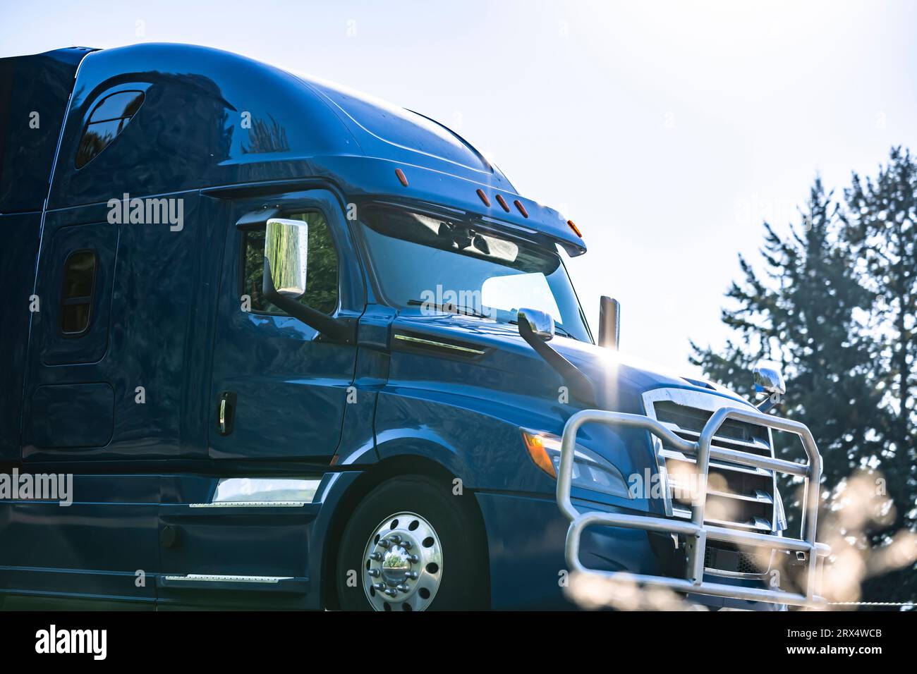 Blue industrial carrier big rig semi truck tractor with heavy duty pipes grille guard and glare from the sun rays on the body running on the highway r Stock Photo