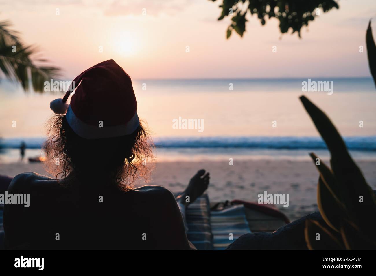 Man with backlit hair and santa hat enjoying Christmas on tropical beach and enjoys the view of the sunset over de ocean, Thailand Stock Photo