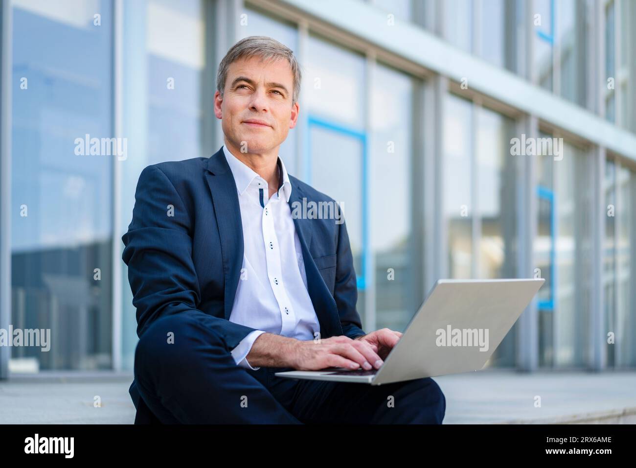 Businessman working on laptop sitting in front of office building Stock Photo