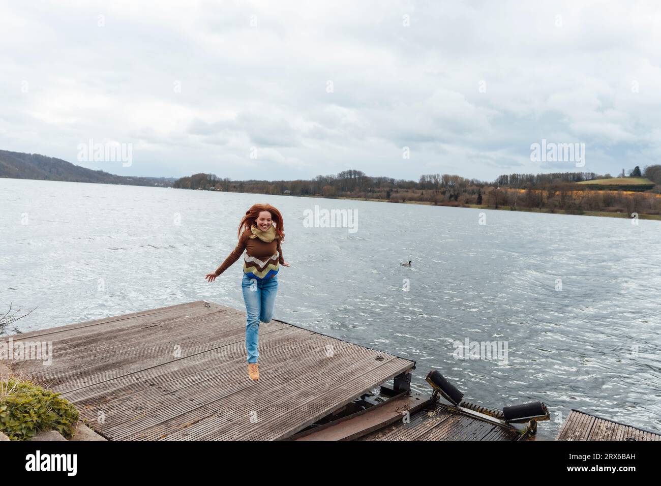 Carefree woman jumping on pier by lake under cloudy sky Stock Photo