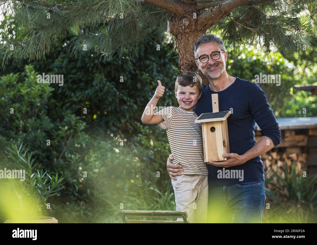 Smiling boy showing thumbs up with grandfather holding birdhouse Stock Photo