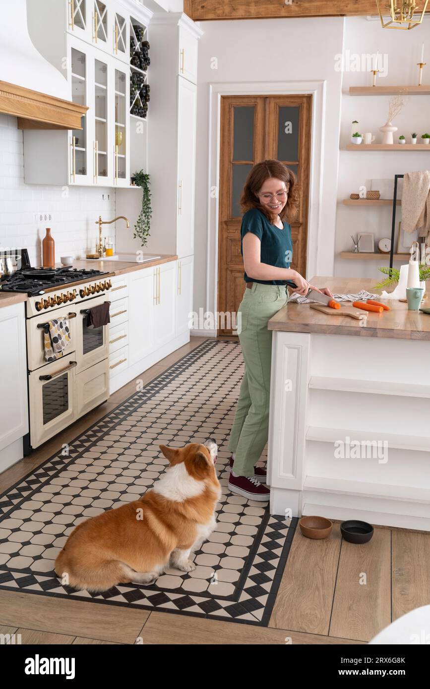 Woman cutting vegetable looking at dog in kitchen Stock Photo