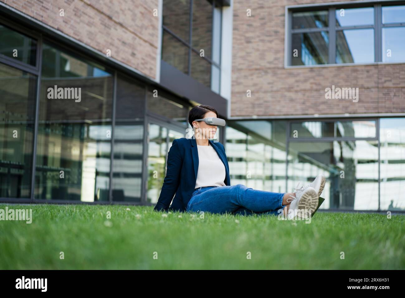 Businesswoman with VR headset sitting on grass at office park Stock Photo