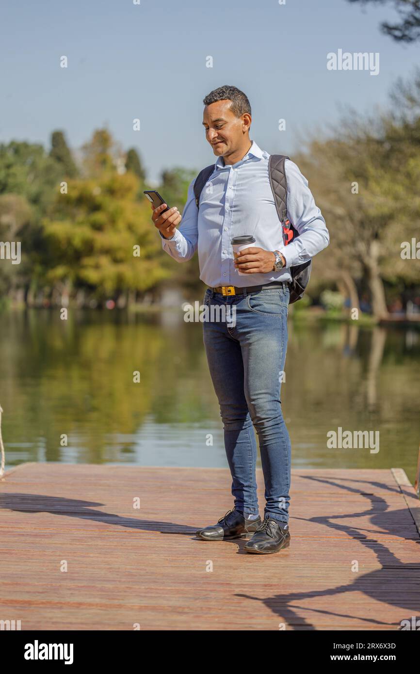 Latin man on a pier with a disposable cup of coffee in his hand looking at his mobile phone. Stock Photo