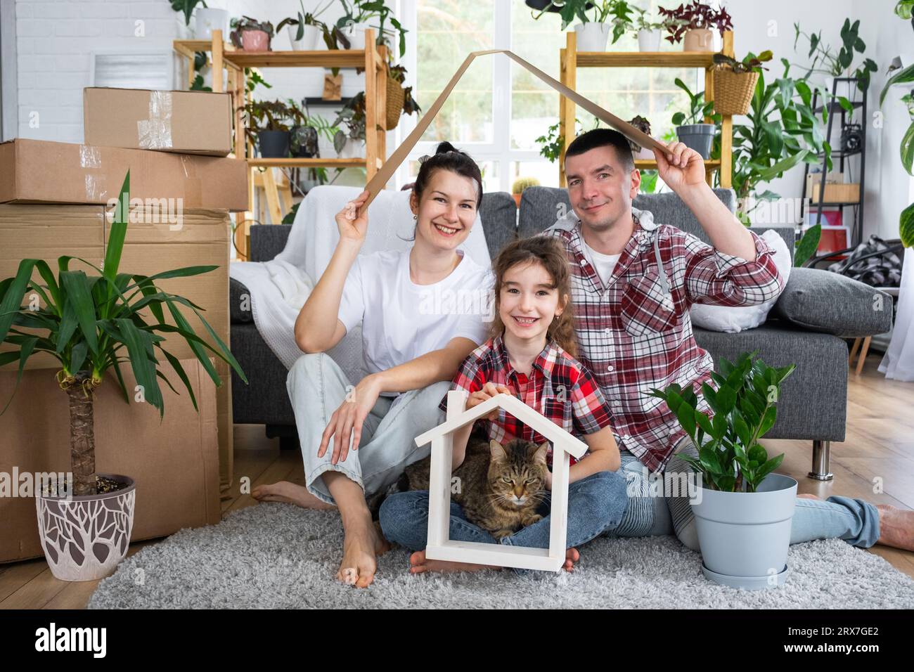 Moved family with a child and a cat pet are enjoying a new home, sitting on the sofa and holding the roof near boxes with things. Mortgage, insurance, Stock Photo