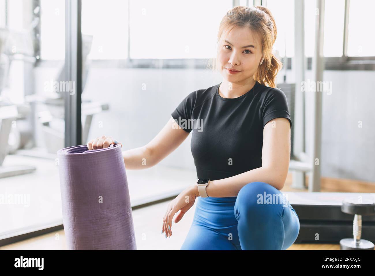 Fitness woman in sport club studio with yoga mat. Portrait happy young woman hand holding pilates mat roll at a fitness training class. Stock Photo