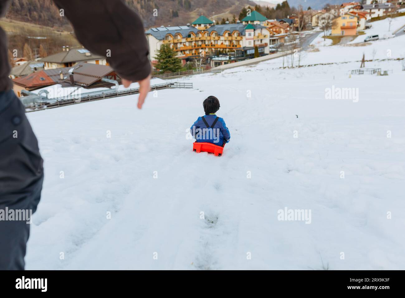 wide angle view of parent helping child with short dark hair to slide down the snowy hill Stock Photo