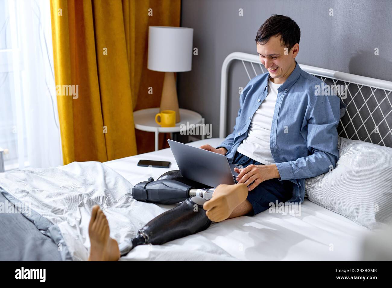 positive male with disability and special needs sit on bed and work on laptop. cheerful and excited. Handicapped Caucasian male using laptop for work Stock Photo