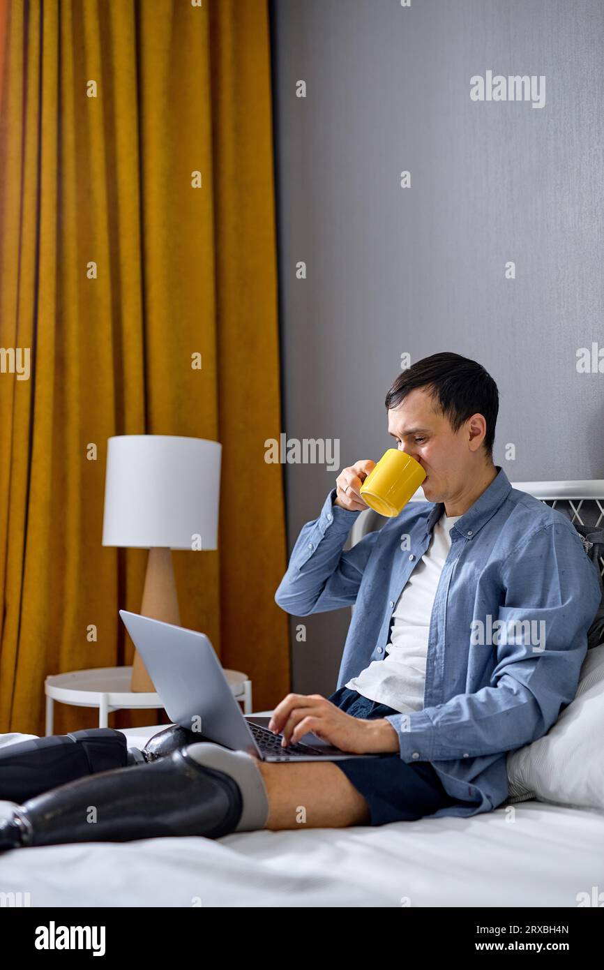 young man with prosthesis on legs using laptop for remote job at home, sitting on bed and drinking tea. Confident Caucasian guy with physical disabili Stock Photo