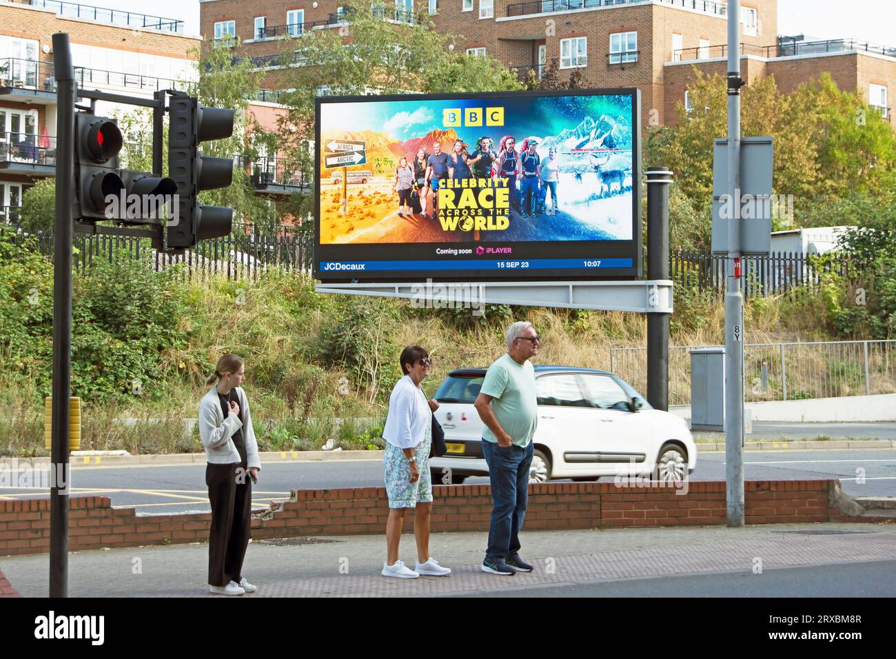 pedestrians wait to cross the road in front of a digital billboard advertising a bbc television programme, celebrity race across the world Stock Photo