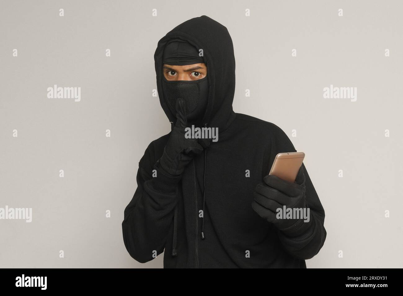 Portrait of mysterious man wearing black hoodie and mask doing hacking activity on mobile phone, hacker holding a smartphone. Isolated image on gray b Stock Photo