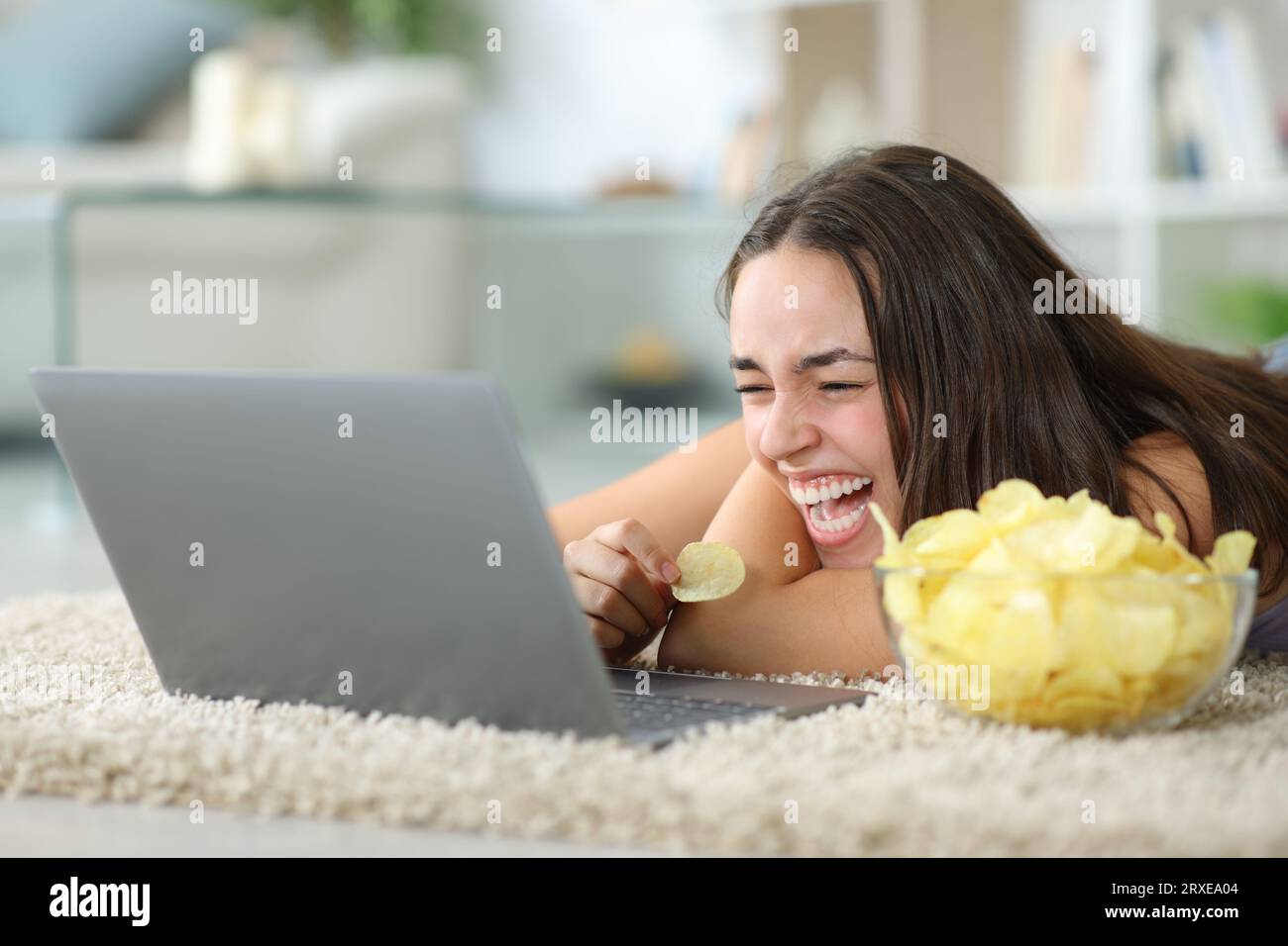 Funny woman watching media on laptop eating chips laughing hilariously at home Stock Photo