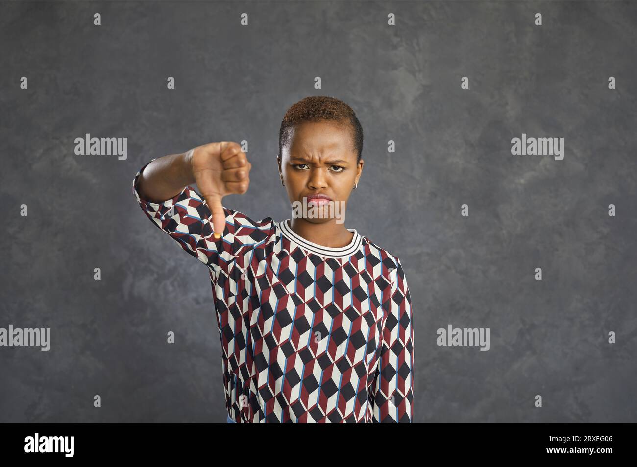 Annoyed woman showing thumbs down while standing with frowning face on gray background. Stock Photo