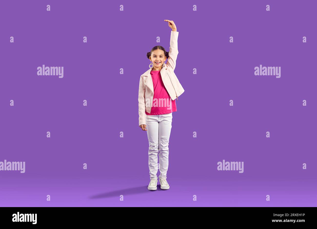 Funny little kid girl hold hand above head showing how to increase her height on purple background. Stock Photo