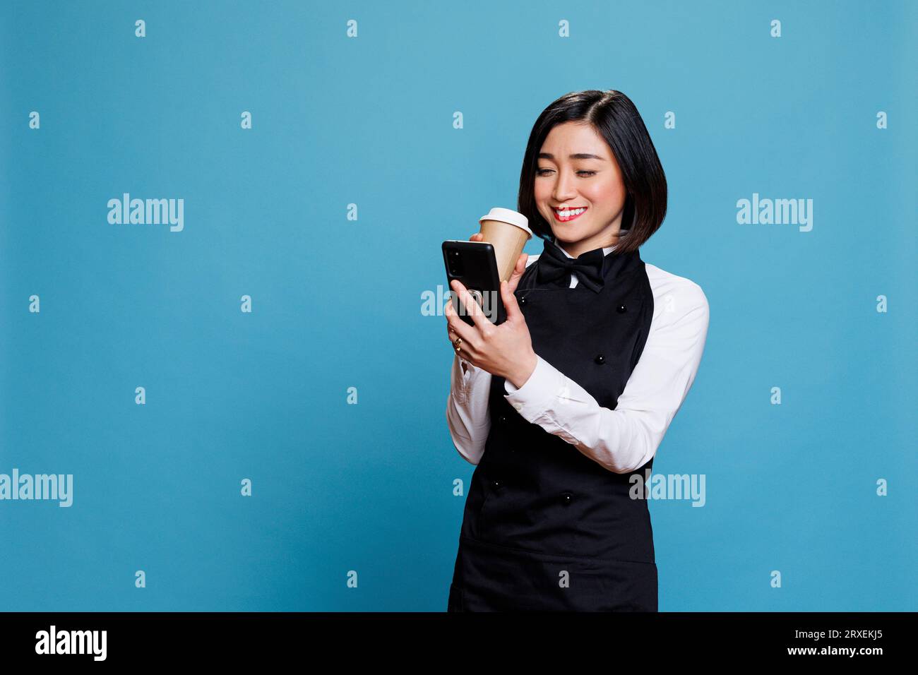 Smiling asian waitress wearing cafe uniform enjoying coffee to go and texting on smartphone. Cheerful woman receptionist holding tea paper mug and reading message on mobile phone Stock Photo