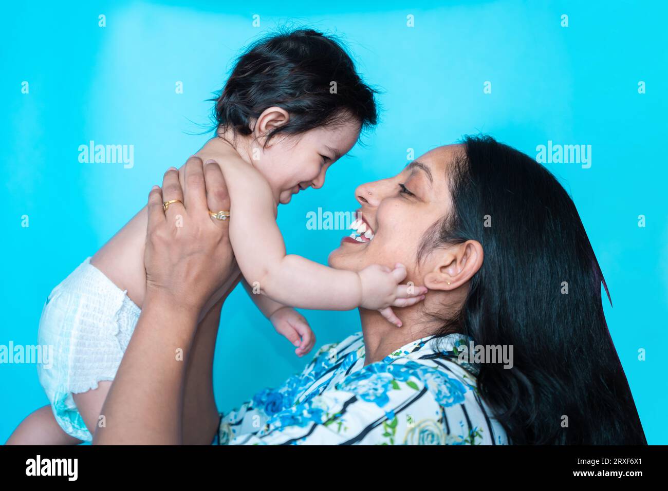 Smiling indian mother playing with her six months cute little baby in diaper isolated over blue background. Happy family. Asian mom with infant child Stock Photo